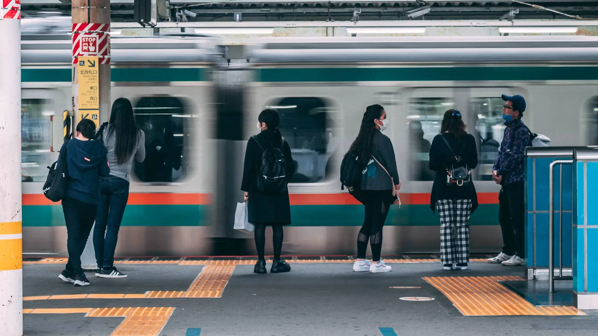 People stand on a subway platform watching a passing silver train, many wearing masks and backpacks nearby.
