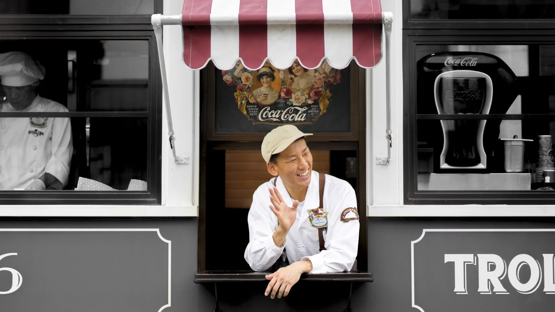 Food truck vendor waves from the service window, wearing a beige cap and white uniform under a red-and-white striped awning with a Coca‑Cola poster behind him.
